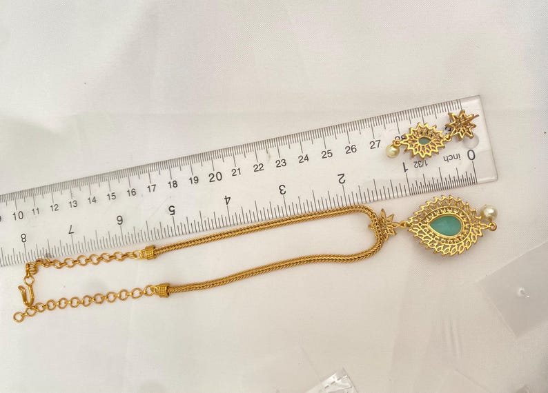 May include: Gold-toned necklace and earrings set. The necklace features a teardrop pendant with a teal-colored stone and a delicate chain. The earrings have a similar design, with a starburst pattern and a small pearl. A ruler is placed next to the jewelry.