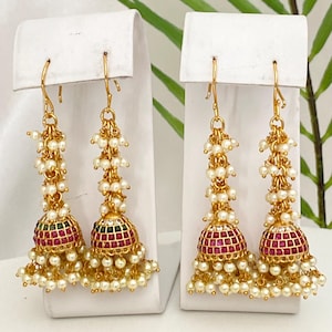 May include: A pair of gold-toned earrings with a bell-shaped design. The earrings feature a red and green patterned top, adorned with small white pearls and a dangling cluster of pearls. The earrings are displayed on a white stand.