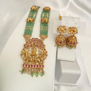 May include: A gold-toned necklace and earring set. The necklace features multiple strands of green beads, gold elephant accents, and a pendant with red and gold details. The earrings have a similar design with gold elephants and red accents. The set is displayed on a white surface.