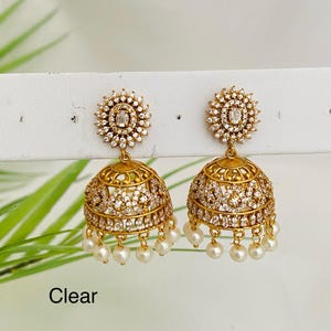 May include: Gold-toned jhumka earrings with clear crystals and dangling pearls. The earrings have a floral design at the top and a bell-shaped bottom. The word "Clear" is at the bottom of the image.