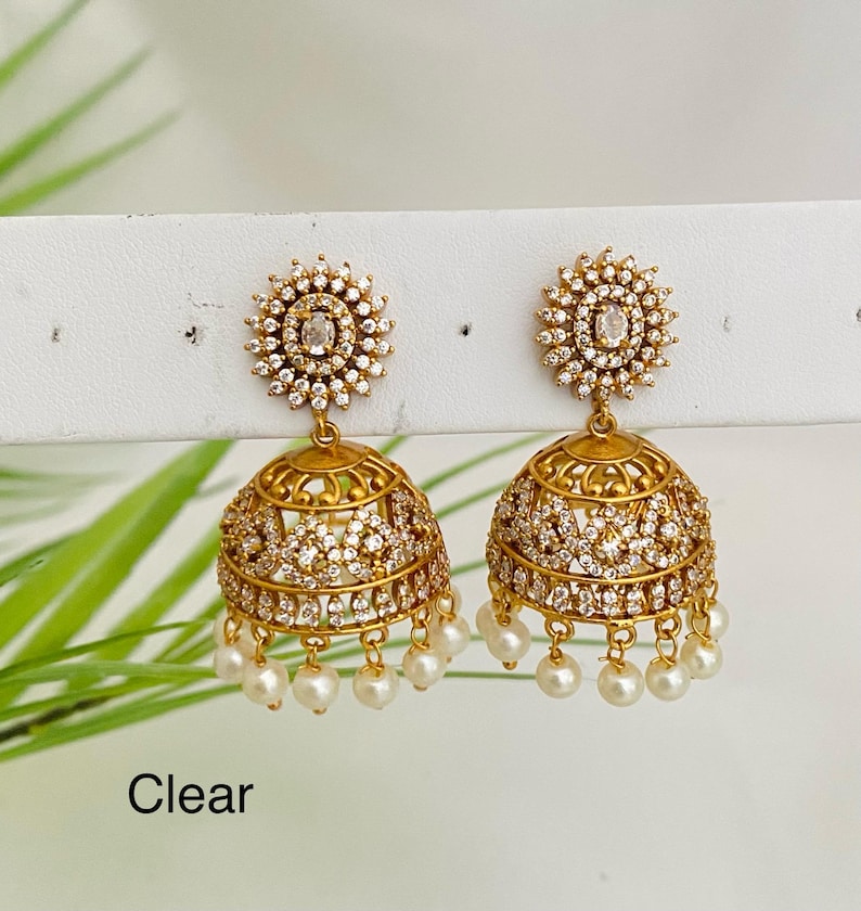 May include: Gold-toned jhumka earrings with clear crystals and dangling pearls. The earrings have a floral design at the top and a bell-shaped bottom. The word "Clear" is at the bottom of the image.