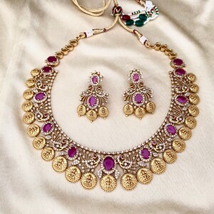 May include: A gold necklace with a coin design and red gemstones. The necklace is paired with matching earrings with red gemstones.