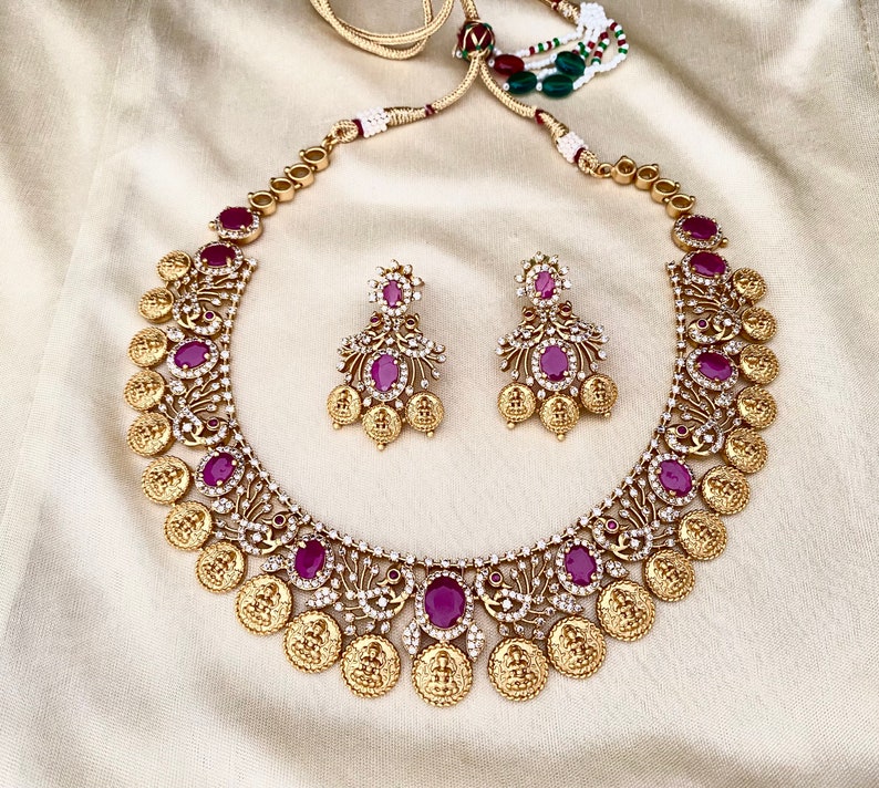 May include: A gold necklace with a coin design and red gemstones. The necklace is paired with matching earrings with red gemstones.