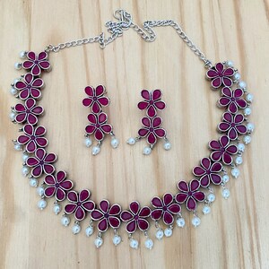 Op de afbeelding: Een zilveren ketting met een roze bloemenontwerp en parel accenten. Passende oorbellen met roze bloemen en parels.
