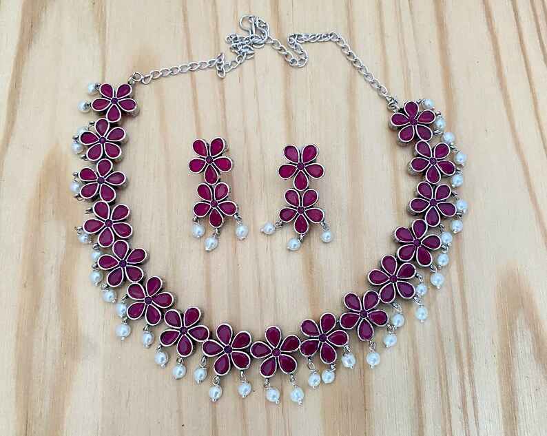 Op de afbeelding: Een zilveren ketting met een roze bloemenontwerp en parel accenten. Passende oorbellen met roze bloemen en parels.