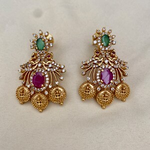 May include: Gold tone earrings with a floral design, featuring a pink and green gemstone. The earrings have a dangling design with multiple gold coins.