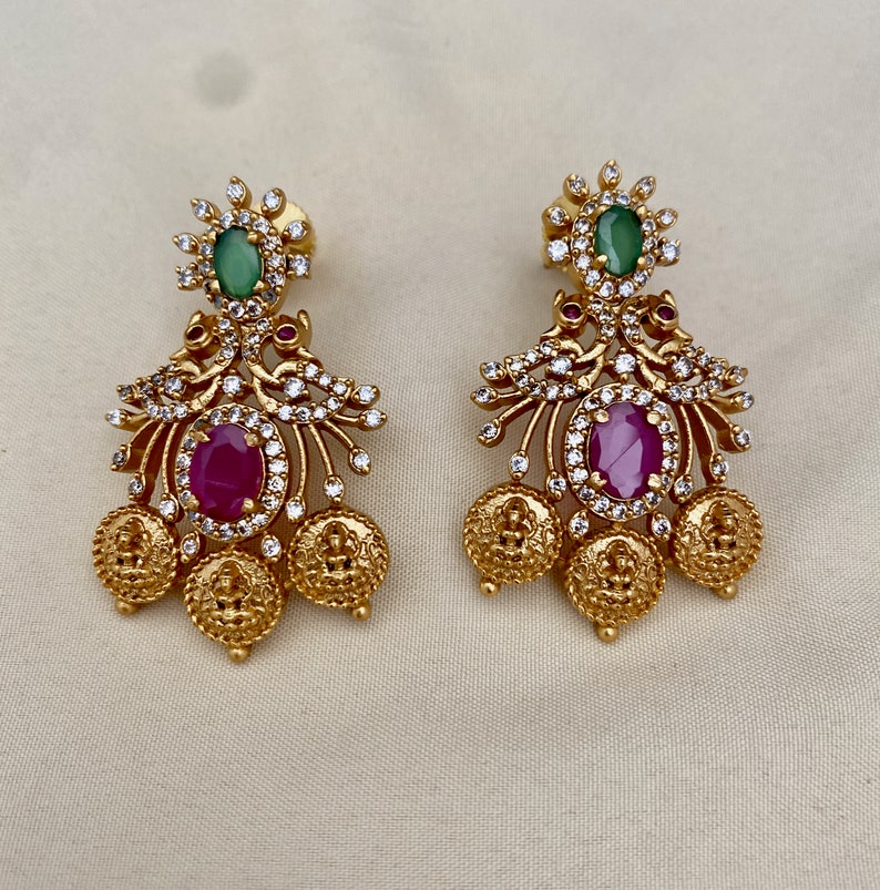 May include: Gold tone earrings with a floral design, featuring a pink and green gemstone. The earrings have a dangling design with multiple gold coins.