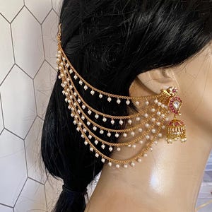 May include: Gold-toned ear cuff with multiple pearl strands and a matching earring. The earring features red and white accents and a bell-shaped charm. The ear cuff attaches to the hair and drapes down the ear.