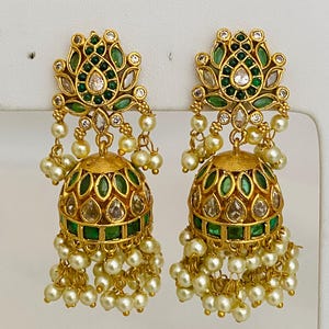 May include: A pair of gold-toned earrings with a floral design. The earrings feature green and clear gemstones, with a dangling cluster of small, round, off-white pearls. The top portion has a lotus-like design, and the bottom is a bell shape.
