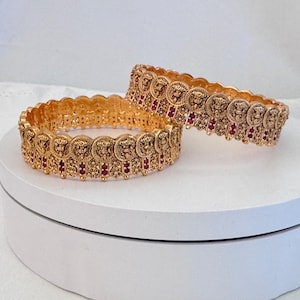 May include: Two gold-toned bangles featuring a repeating circular motif with embossed lion head designs. Each circular element is separated by small vertical bars accented with tiny, deep red stones, creating a textured, ornate appearance.