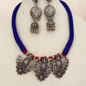 May include: A silver necklace with a blue and red cord and three silver pendants featuring a goddess figure. The necklace is paired with silver dangle earrings with a circular design.