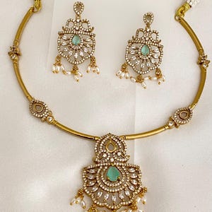 May include: A gold-toned necklace and matching earrings set. The necklace features a pendant with a central teal stone, surrounded by clear crystals and dangling pearls. The earrings mirror the pendant design, creating a cohesive jewelry set.