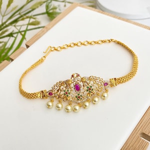 May include: A gold-toned necklace with a central pendant featuring a large, oval, magenta-colored stone, surrounded by smaller clear stones and accents of red and green. The pendant is adorned with dangling faux pearls. The necklace has a chain-like design.