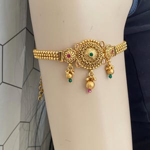 May include: A gold-toned armlet featuring a central circular medallion with intricate filigree patterns, accented by small green and pink stones. The band consists of multiple rows of small gold beads, with three dangling charms featuring beads.