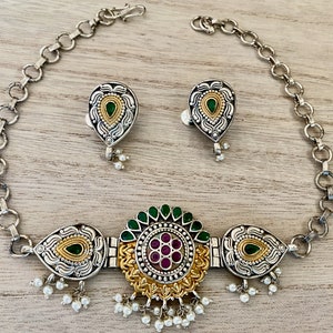 May include: A silver and gold necklace with a green and pink floral design. The necklace has a chain and a pendant with a floral design. The pendant has a green stone in the centre and is surrounded by smaller pink stones. The necklace also has a pair of matching earrings.