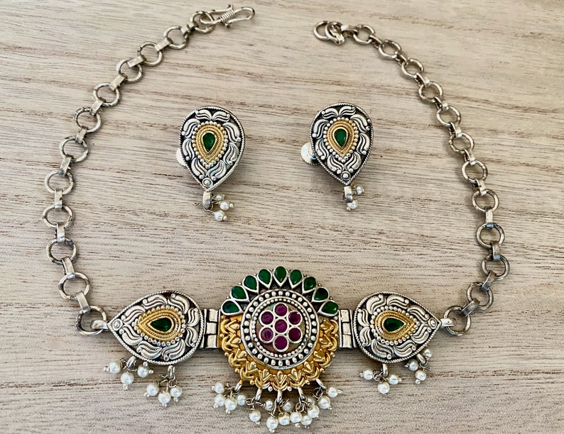 May include: A silver and gold necklace with a green and pink floral design. The necklace has a chain and a pendant with a floral design. The pendant has a green stone in the centre and is surrounded by smaller pink stones. The necklace also has a pair of matching earrings.