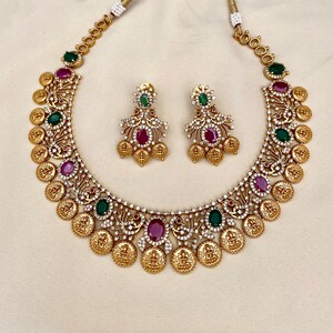 May include: A gold necklace with a coin design and green and red gemstones. Matching earrings with the same design.
