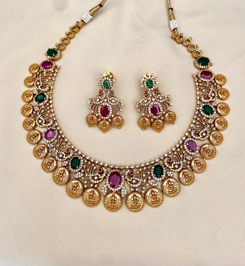 May include: A gold necklace with a coin design and green and red gemstones. Matching earrings with the same design.