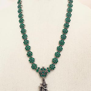 May include: A silver necklace with a green floral design and a pendant featuring a silver figure playing a flute. The pendant is surrounded by green stones and has a fringe of white pearls.