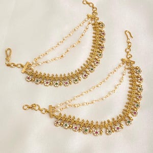 May include: Two gold-toned ear cuffs with a floral design, featuring red, green, and white gemstones. Each cuff has two pearl chains and a hook closure. The jewelry is displayed on a white surface.