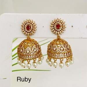 May include: A pair of gold-toned jhumka earrings with a ruby center and white gemstones. The earrings have a bell shape with dangling white pearls. The word "Ruby" is printed at the bottom.