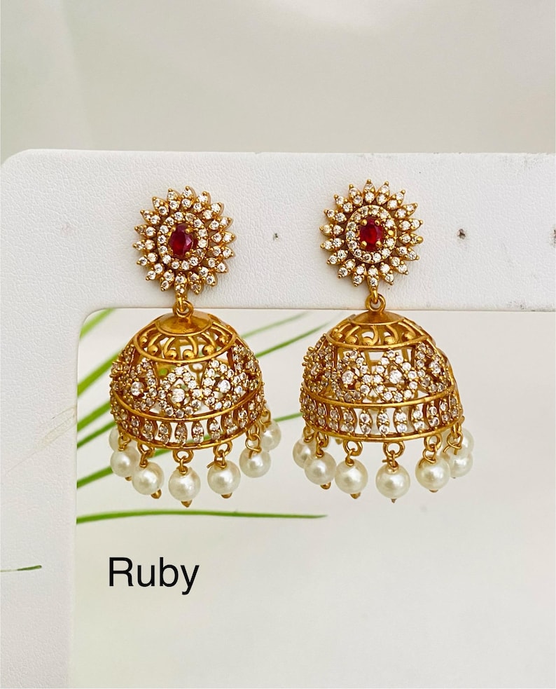 May include: A pair of gold-toned jhumka earrings with a ruby center and white gemstones. The earrings have a bell shape with dangling white pearls. The word "Ruby" is printed at the bottom.