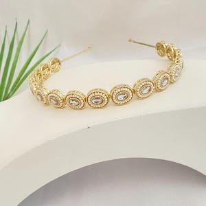 May include: A gold-toned headband with oval-shaped, clear crystal accents. The crystals are set in a gold frame and are surrounded by smaller, clear crystals. The headband is displayed on a white surface.