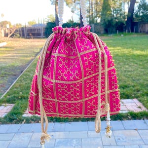 May include: A pink drawstring bag with gold embroidery and a white beaded handle. The bag has a square pattern and is decorated with small gold beads.