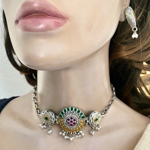 May include: A silver and gold choker necklace with a round pendant featuring green and pink stones. The pendant has a floral design and is surrounded by smaller pearls. The necklace has a chain link design and is accented with small pearl drops.