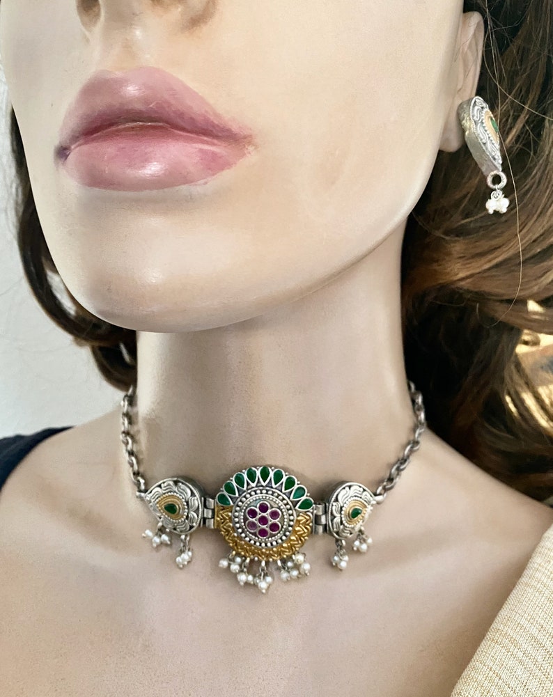 May include: A silver and gold choker necklace with a round pendant featuring green and pink stones. The pendant has a floral design and is surrounded by smaller pearls. The necklace has a chain link design and is accented with small pearl drops.