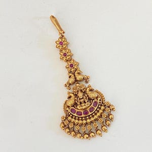 May include: A gold-toned maang tikka headpiece featuring a vertical chain of three flower motifs with embedded red stones, leading to a central pendant adorned with two peacock figures, a deity figure, crescent accents, and dangling beads.