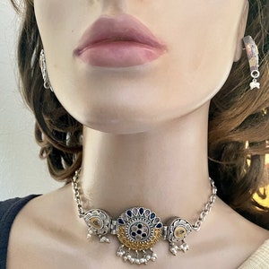 May include: A silver and gold choker necklace with a large, round pendant featuring blue and pink stones. The pendant is surrounded by intricate floral designs. The necklace has a chain link design and is adorned with small pearl-like beads.