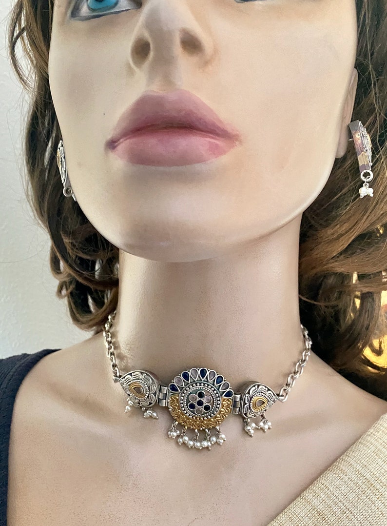 May include: A silver and gold choker necklace with a large, round pendant featuring blue and pink stones. The pendant is surrounded by intricate floral designs. The necklace has a chain link design and is adorned with small pearl-like beads.