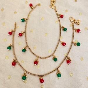 May include: A pair of gold anklets with red and green beads. The anklets are made of a delicate chain and have a dangling design.