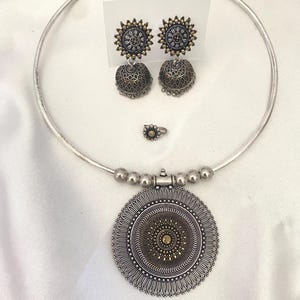 May include: A silver-toned jewelry set consisting of a rigid choker necklace with a large circular pendant, a pair of matching jhumka earrings with sunburst tops, and a small matching ring. The pieces feature intricate oxidized metal detailing.