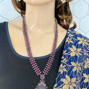 May include: A silver necklace with a pink stone pendant and matching earrings. The necklace features a long chain with pink stones and a large pendant with a floral design. The earrings are dangle earrings with a similar design to the pendant.