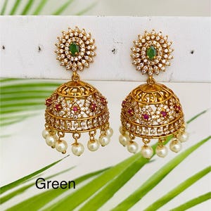 May include: A pair of gold-toned earrings with a floral design. Each earring features a green gemstone center surrounded by clear crystals. The lower portion has a bell shape with red gemstones and dangling faux pearls. The word "Green" is visible.