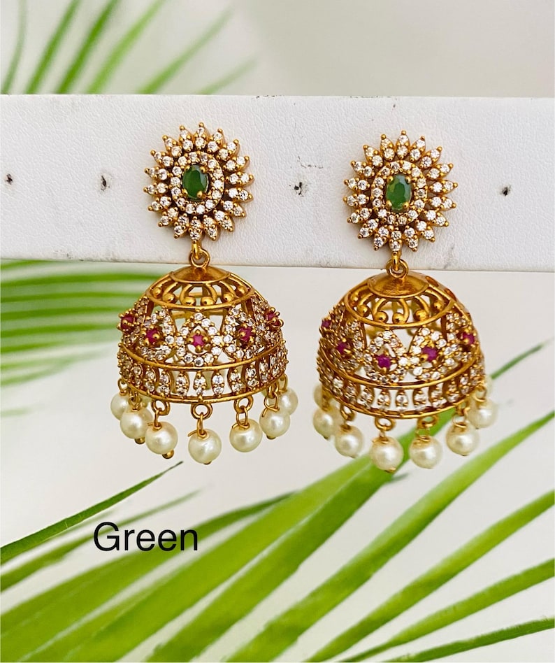 May include: A pair of gold-toned earrings with a floral design. Each earring features a green gemstone center surrounded by clear crystals. The lower portion has a bell shape with red gemstones and dangling faux pearls. The word "Green" is visible.