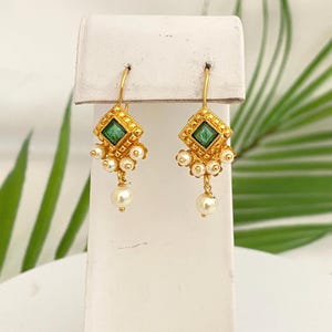May include: A pair of gold-colored earrings with a square, emerald-green center stone. The earrings have a diamond-shaped design with small white pearls dangling below. The earrings are displayed on a white stand.
