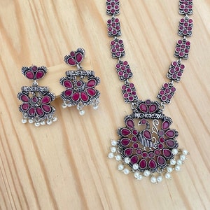 May include: A silver necklace with a pink stone peacock pendant and matching earrings. The necklace is made of a chain of square-shaped links with pink stones. The pendant features a peacock design with pink stones and a pearl drop.