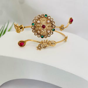 May include: Gold-toned arm cuff with a central circular design featuring a red center stone, surrounded by clear and green stones. The cuff has ornate floral and leaf details, with red teardrop-shaped accents on each end.