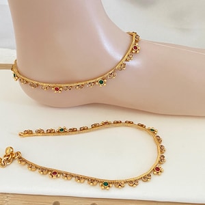 May include: A pair of gold-toned anklets featuring a twisted chain design adorned with small, alternating red and green circular accents along the length. One anklet is displayed on a foot model, while the other lies flat, showing a small bell charm.