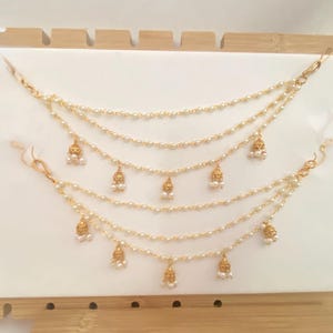 May include: A multi-layered gold-toned chain necklace featuring small white beads along the strands and dangling bell-shaped charms with tiny white bead accents. The jewelry piece is displayed on a white board with gold-toned metal clasps at the ends.