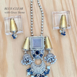 May include: A silver and gold necklace with a gray stone pendant, blue and white beads, and two matching earrings. The necklace has a chain with small silver beads. The pendant has a square frame with a gray stone in the center, surrounded by smaller blue and white stones. The earrings are similar to the pendant, with a smaller version of the square frame and gray stone.