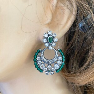 May include: Silver earrings with a green stone and white stones in a floral design. The earrings are surrounded by green beads.