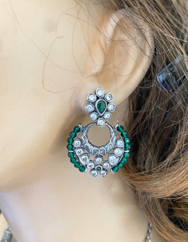 May include: Silver earrings with a green stone and white stones in a floral design. The earrings are surrounded by green beads.