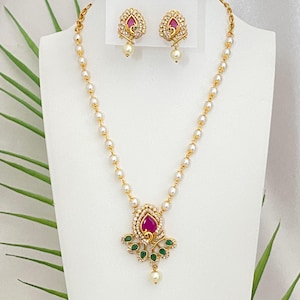 May include: A gold-toned necklace and earring set. The necklace features a pendant with a teardrop-shaped, ruby-colored stone, surrounded by clear crystals and green accents. The earrings mirror the pendant design, with a pearl drop.