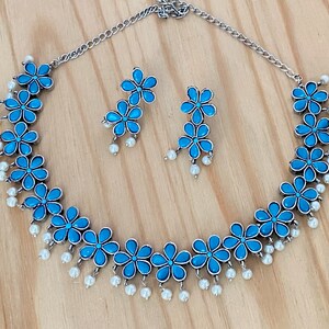 Op de afbeelding: Een zilveren ketting en oorbellen set met blauwe bloemenaccenten en witte parel druppels.