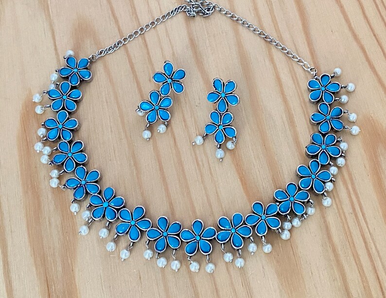 Op de afbeelding: Een zilveren ketting en oorbellen set met blauwe bloemenaccenten en witte parel druppels.