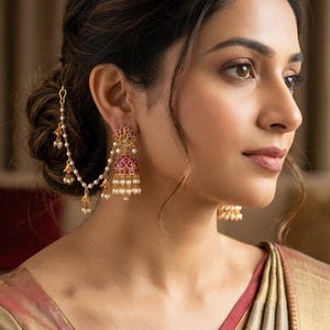 May include: A gold-toned jhumka earring featuring a bell-shaped pendant inlaid with small red stones and accented with white pearls. A delicate gold chain connects the earring to a hair pin, adorned with additional pearls and small gold charms.